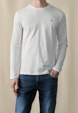 Langärmliges weißes T-Shirt aus Baumwolle; verfügt über einen Rundhalsausschnitt und ein kleines schwarzes Logo auf der Brust. Getragen mit dunkelblauen Jeans.