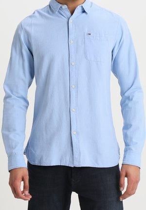 Mann trägt ein hellblaues, langärmliges Hemd mit Knöpfen und einer Brusttasche, kombiniert mit dunklen Jeans, vor einem schlichten Hintergrund.