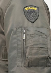 Grå jakke med et fremtrædende sort og gult "Blauer" mærke, lynlåslomme og tekstiler med detaljer i syninger.