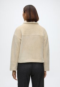 Šviesiai rudos vilnos striukė su trumpu dizainu, plačiomis rankovėmis ir klasikiniu kaklu. Pasižymi tekstūruotu paviršiumi ir siūlės detale nugaroje.