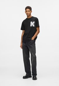 Jauns vīrietis stāv ar roku kabatā, ģērbies melnās grafiskās T-kreklā, tumšās džinsās un melnās kurpēs pret vienkāršu baltu fonu.