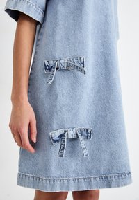 Svetlo modra denim obleka s kratkimi rokavi, ki ima dva naglavna dodatka in ravno rob. Tekstura je gladka z diskretnimi šivi.