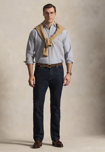 Homme portant des lunettes, chemise bleu clair à boutons avec manches retroussées, pull beige sur les épaules, jean foncé et mocassins marron, debout.