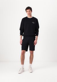 Black sweatshirt with ribbed cuffs, paired with dark shorts. White sneakers and socks complete the outfit, set against a plain background.