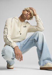 Beige kapucnis pulóver fekete díszítéssel és Puma logóval, világoskék, laza szabású farmernadrággal és krémszínű sportcipőkkel, barna talppal párosítva.