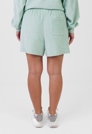 Persona che indossa pantaloncini in spugna verde chiaro con elastico in vita e tasca posteriore, abbinati a una felpa coordinata e sneakers bianche.