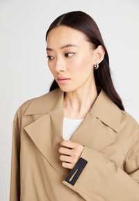 Beige színű trench coat széles gallérral és rejtett gombzárral, fekete téglalap alakú kiegészítővel a mandzsettán. A modellen fehér felső van.