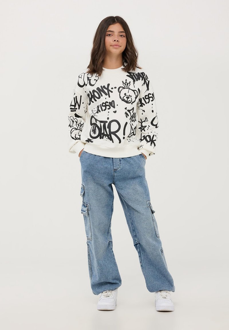 Biała bluza z kapturem z czarnym nadrukiem w stylu graffiti, przedstawiająca niedźwiedzia i tekst. Połączona z jasnoniebieskimi spodniami cargo i białymi trampkami.