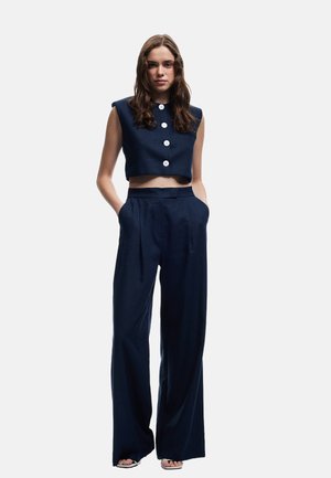Marineblå cropped top uden ærmer med hvide knapper, parret med højtaljede bukser i marineblå med vide ben. Glat stof, minimalistisk design, skræddersyet pasform.