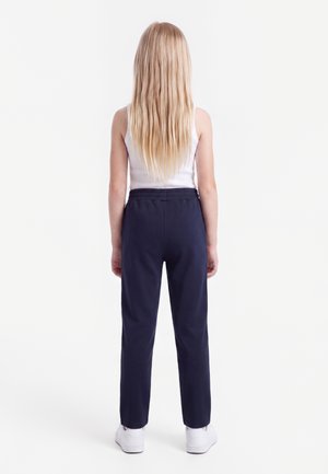 Kind mit langen blonden Haaren steht mit dem Rücken zur Kamera, trägt ein weißes Tanktop, dunkelblaue Hosen mit elastischem Bund und weiße Schuhe.
