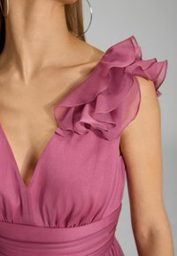 Robe fuchsia avec un décolleté en V profond, ornée d'accents en tulle plissé sur les épaules et d'une taille froncée. Texture lisse dans l'ensemble.