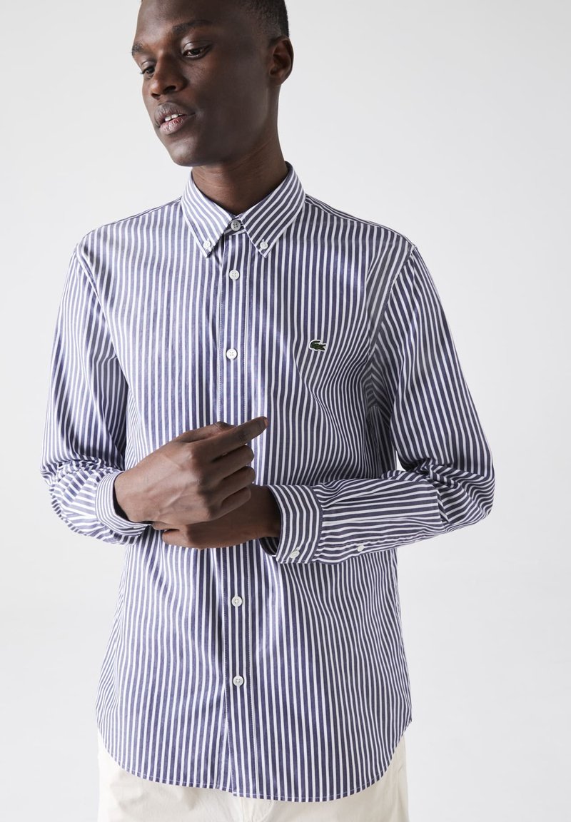 Button-up shirt in blue and white stripes, featuring a collar, long sleeves, and a small green logo. Smooth cotton fabric, traditional fit.
