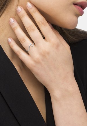 Anello d'argento a forma di onda indossato su una mano, con una texture liscia e lucida. La mano mostra unghie naturali ed è su uno sfondo nero.