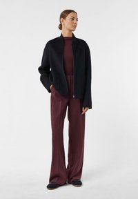Schwarze Zip-Jacke mit Stehkragen, kombiniert mit weiten burgunderfarbenen Hosen. Der Stoff wirkt glatt, und die Jacke hat Taschen.