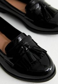Mocasines de charol negro con acentos de borlas, que cuentan con una punta redondeada, un diseño elegante y una suela texturizada.