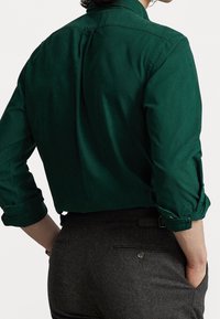 Ciemnozielona koszula na guziki z podwiniętymi rękawami i plisą z tyłu; zestawiona z ciemnoszarymi, dopasowanymi spodniami z tylną kieszenią.