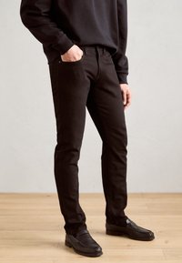 Vaqueros de denim negro con corte ajustado, que presentan un clásico diseño de cinco bolsillos y costuras sutiles. Llevados con mocasines negros y un sudadera.