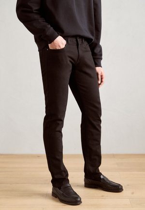 Jean en denim noir avec une coupe slim, présentant un design classique à cinq poches et une couture subtile. Porté avec des mocassins noirs et un sweat-shirt.