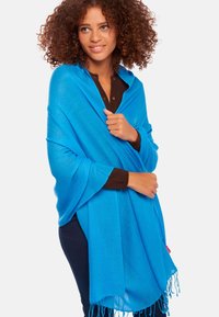 Châle bleu en tissu doux et texturé, doté d'un ourlet à franges et d'un design drapé. Porté sur un haut marron foncé.