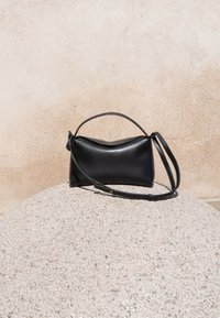 Sac à main en cuir noir avec une poignée courte et une longue bandoulière réglable, posé sur une surface ronde en pierre texturée contre un mur beige.
