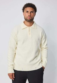 Pull en tricot couleur crème avec un col, trois boutons, des poignets et un ourlet côtelés, et une texture douce. Porté avec un pantalon noir.