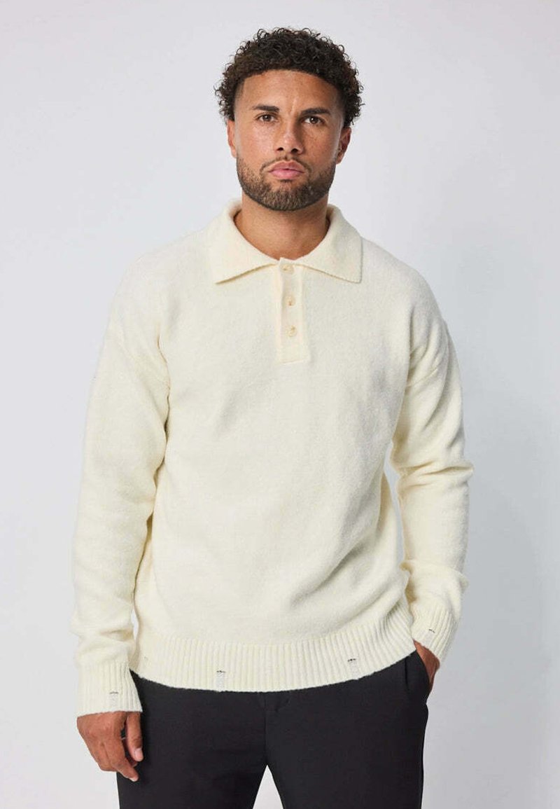 Pull en tricot couleur crème avec un col, trois boutons, des poignets et un ourlet côtelés, et une texture douce. Porté avec un pantalon noir.