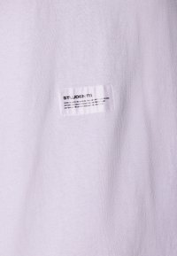 Hvit bomull t-skjorte med et rektangulært merke med svart tekst. Stoffet har en jevn tekstur og et enkelt, rent design.