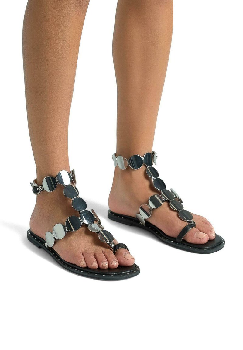 Černé ploché sandály s kulatými kovovými akcenty uspořádanými v dekorativním vzoru, se zapínáním T-strap a hřebíčkovým detailem podél podrážky.