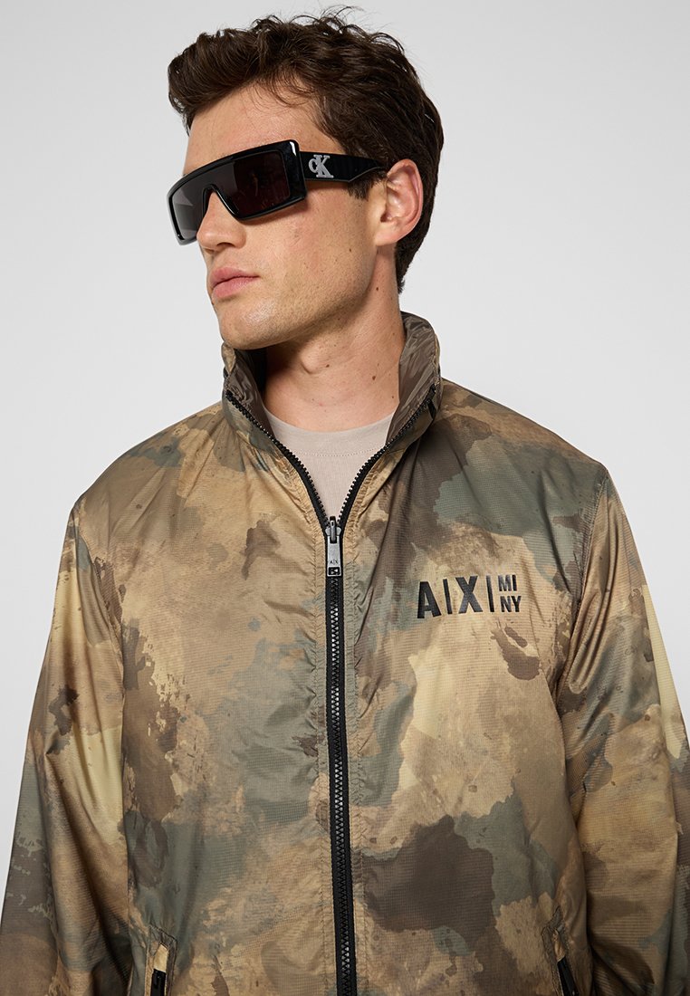 M baconouflage-jasje, lichtgewicht met een hoge kraag en ritssluiting. Beschikt over een logo op de borst en een gladde textuur.