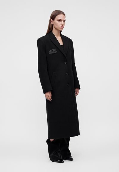 Femme portant un long manteau noir sur mesure à trois boutons, debout avec une posture droite sur un fond clair uni.