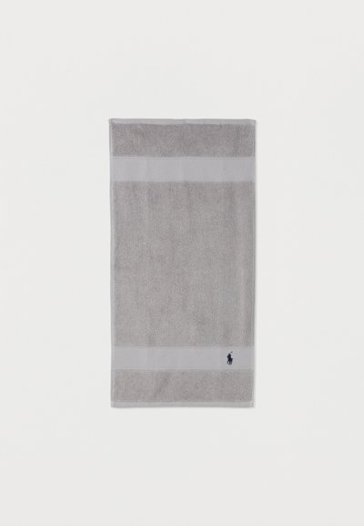 Πετσέτα ορθογώνια σε ανοιχτό γκρι χρώμα με υφασμένες λωρίδες κοντά στις άκρες και μικρό κεντητό λογότυπο σε σκούρο μπλε στο κάτω δεξιό μέρος.