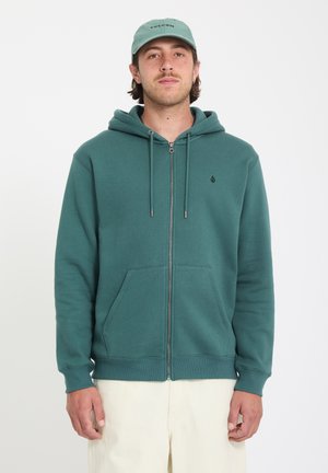 Mies, joka pukeutunut teal-väriseen vetoketjulliseen huppariin ja vaaleanvihreään lippalakkiin, seisoo yksinkertaisen valkoisen taustan edessä.