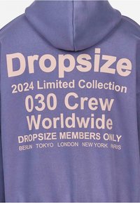 Heled lilla kapuutsilm, millel on suur valge kiri. Kiri sisaldab sõnu "Dropsize," "2024 Limited Collection" ja linnade nimesid: Berliin, Tokyo, London, New York, Pariis.