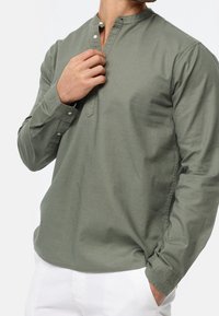 Camisa de manga larga color verde oliva con cuello mandarín, detalles de botones y tela suave y texturizada. Combinada con pantalones blancos.