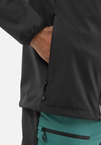 Veste softshell noire avec poches zippées, poignets élastiques et panneaux teal contrastants au bas et aux poches. Tissu lisse et durable.
