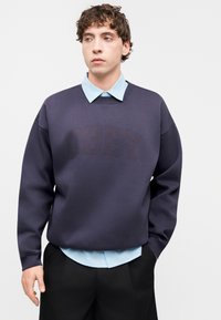 Σκούρο μπλε φούτερ με την επιγραφή «OBEY» σε διακριτικό τόνο, φορεμένο πάνω από ανοιχτό μπλε πουκάμισο με γιακά, συνδυασμένο με μαύρο παντελόνι. Μαλακή υφή.