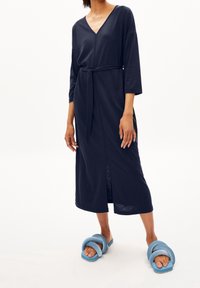 Granatowa sukienka o długim rękawie, zapinana w talii, z dekoltem w kształcie litery V; ma boczne rozcięcie, gładką fakturę i luźny krój. Noszona z niebieskimi sandałami.
