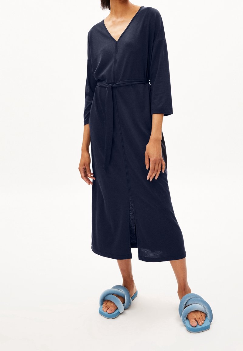Granatowa sukienka o długim rękawie, zapinana w talii, z dekoltem w kształcie litery V; ma boczne rozcięcie, gładką fakturę i luźny krój. Noszona z niebieskimi sandałami.