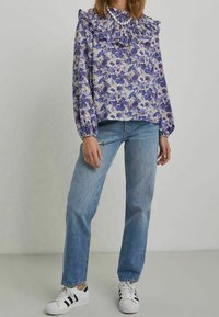 Blommig blus i lila och vitt med volangkrage, långa ärmar, och matchad med ljusblå jeans och svart-randiga vita sneakers.