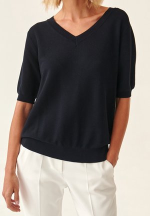 Kvinde iført en sort kortærmet V-hals sweater og hvide bukser med højtaljet, med den ene hånd i lommen mod en ensfarvet baggrund.