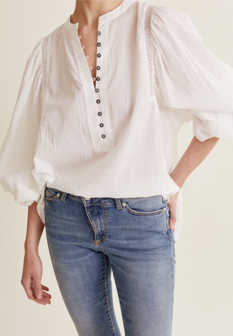 Femme portant une blouse blanche avec des boutons noirs et des détails en dentelle, associée à un jean bleu denim, debout avec une main dans la poche.
