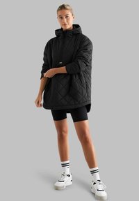 Zwarte gewatteerde oversized hoodie met een ritssluiting, een capuchon en korte mouwen. Gekozen boven zwarte fietsshorts en gecombineerd met witte sneakers.