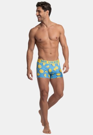 Uomo sorridente senza maglietta che cammina a piedi nudi indossando boxer blu con motivi di limoni gialli e una fascia gialla con la scritta "MR UKKO."
