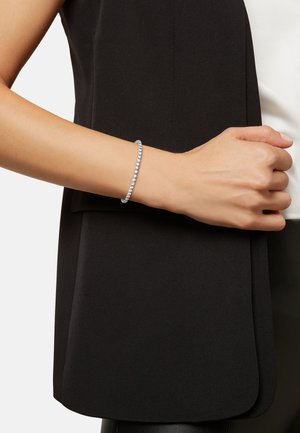 Pulsera de plata con piedras redondas y transparentes engastadas en una banda flexible, llevada en una muñeca sobre un fondo de ropa negra.