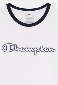 Tricou alb din bumbac, cu guler rotund ribbed în nuanță de bleumarin și un mare logo brodat "Champion" în albastru închis, având un efect de umbră.