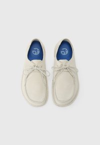 Pantofi din suede bej cu vârful rotunjit și design cu șireturi. Interiorul prezintă o branț albastru cu un logo. Cusăturile sunt vizibile de-a lungul perimetrului.