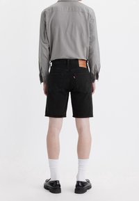 Shorts de mezclilla negra con corte recto, que cuentan con dos bolsillos traseros y un parche de cuero en la cintura, combinados con una camisa de manga larga gris.