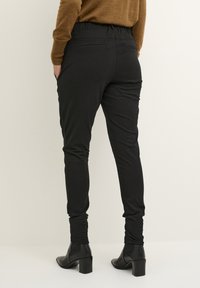 Pantalons noirs ajustés avec une taille élastique, deux poches latérales et une conception de jambe fuselée. Portés avec des bottes à talons noires arrivant aux chevilles.