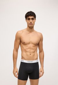 Fekete boxeralsó geometrikus mintával, fehér derékszárral, amelyen a "Calvin Klein" márkázás található, testhezálló fazonban, sima anyagú textúrával.