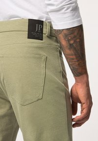 Pantalons vert clair avec une finition texturée ; dotés d'une poche arrière, d'une étiquette à la taille indiquant "JP 1880", et d'un tatouage partiellement visible sur le bras.
